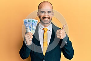 Young hispanic business man holding south african 100 rand banknotes smiling happy and positive, thumb up doing excellent and