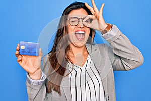 Young hispanic business woman holding credit card over blue isolated background with happy face smiling doing ok sign with hand on