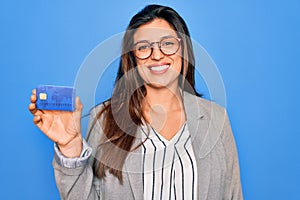 Young hispanic business woman holding credit card over blue isolated background with a happy face standing and smiling with a