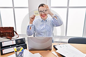 Young hispanic business woman at the office calculating taxes smiling happy doing ok sign with hand on eye looking through fingers