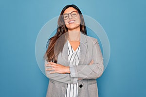 Young hispanic business woman wearing glasses standing over blue isolated background happy face smiling with crossed arms looking
