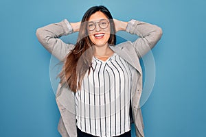 Young hispanic business woman wearing glasses standing over blue isolated background relaxing and stretching, arms and hands