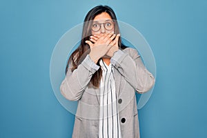 Young hispanic business woman wearing glasses standing over blue isolated background shocked covering mouth with hands for mistake