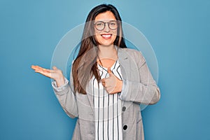 Young hispanic business woman wearing glasses standing over blue isolated background Showing palm hand and doing ok gesture with
