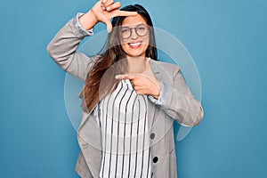 Young hispanic business woman wearing glasses standing over blue isolated background smiling making frame with hands and fingers