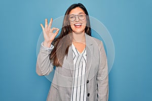 Young hispanic business woman wearing glasses standing over blue isolated background smiling positive doing ok sign with hand and