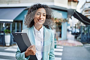 Young hispanic business woman wearing professional look smiling confident at the city holding worker clipboard