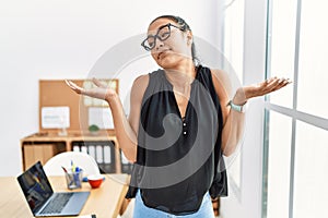 Young hispanic business woman working at the office clueless and confused expression with arms and hands raised