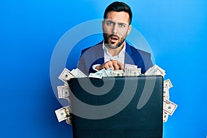 Young hispanic businessman holding briefcase full of dollars in shock face, looking skeptical and sarcastic, surprised with open