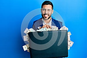 Young hispanic businessman holding briefcase full of dollars sticking tongue out happy with funny expression