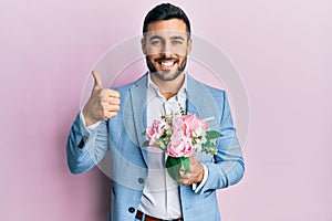 Young hispanic businessman wearing business jacket holding flowers smiling happy and positive, thumb up doing excellent and