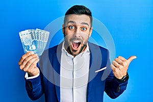 Young hispanic businessman wearing business suit holding chilean pesos banknotes pointing thumb up to the side smiling happy with