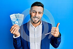 Young hispanic businessman wearing business suit holding chilean pesos banknotes smiling happy and positive, thumb up doing