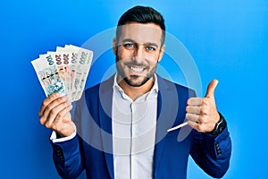 Young hispanic businessman wearing business suit holding czech koruna banknotes smiling happy and positive, thumb up doing