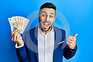 Young hispanic businessman wearing business suit holding denmark krone banknotes pointing thumb up to the side smiling happy with