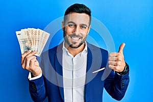 Young hispanic businessman wearing business suit holding denmark krone banknotes smiling happy and positive, thumb up doing