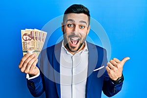 Young hispanic businessman wearing business suit holding hungarian forint banknotes pointing thumb up to the side smiling happy