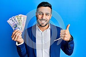 Young hispanic businessman wearing business suit holding indian rupee banknotes smiling happy and positive, thumb up doing