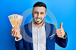 Young hispanic businessman wearing business suit holding norwegian krone banknotes smiling happy and positive, thumb up doing