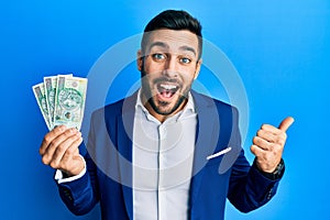 Young hispanic businessman wearing business suit holding polish zloty banknotes pointing thumb up to the side smiling happy with