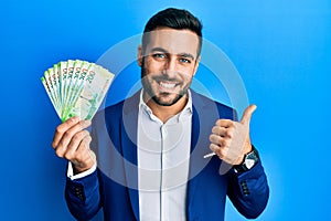 Young hispanic businessman wearing business suit holding russian ruble banknotes smiling happy and positive, thumb up doing