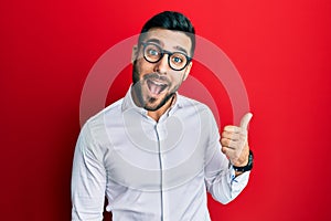 Young hispanic businessman wearing shirt and glasses smiling with happy face looking and pointing to the side with thumb up