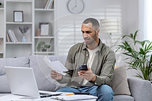 Young hispanic businessman working remotely at home on laptop. Holding the phone and anxiously looking at documents