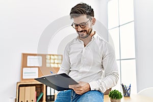 Young hispanic businessman writing on  clipboard sitting on the table at the office