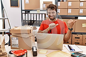 Young hispanic call center agent man working at warehouse pointing to the back behind with hand and thumbs up, smiling confident