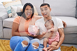 Young hispanic couple with baby holding piggy bank celebrating crazy and amazed for success with open eyes screaming excited