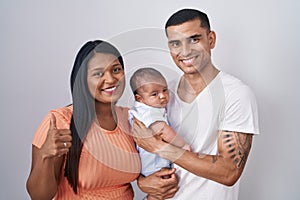 Young hispanic couple with baby standing together over isolated background doing happy thumbs up gesture with hand