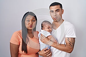 Young hispanic couple with baby standing together over isolated background puffing cheeks with funny face