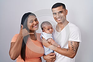 Young hispanic couple with baby standing together over isolated background smiling doing phone gesture with hand and fingers like