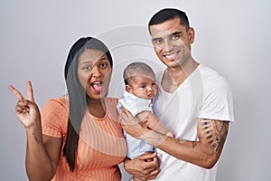Young hispanic couple with baby standing together over isolated background smiling with happy face winking at the camera doing