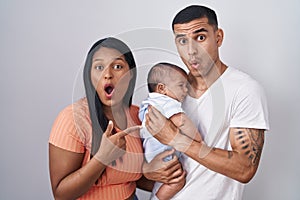 Young hispanic couple with baby standing together over isolated background surprised pointing with finger to the side, open mouth