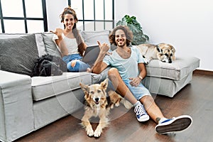 Young hispanic couple with dogs relaxing at home pointing to the back behind with hand and thumbs up, smiling confident