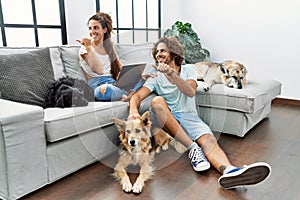 Young hispanic couple with dogs relaxing at home smiling with happy face looking and pointing to the side with thumb up