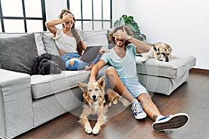 Young hispanic couple with dogs relaxing at home smiling and laughing with hand on face covering eyes for surprise