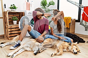 Young hispanic couple doing laundry with dogs covering eyes with arm, looking serious and sad