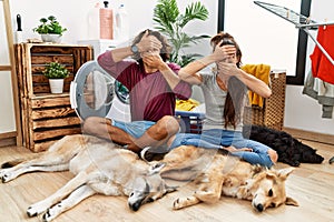 Young hispanic couple doing laundry with dogs covering eyes and mouth with hands, surprised and shocked