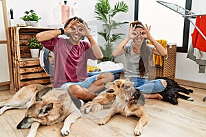 Young hispanic couple doing laundry with dogs doing ok gesture like binoculars sticking tongue out, eyes looking through fingers