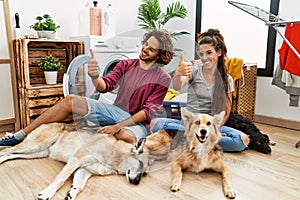 Young hispanic couple doing laundry with dogs looking proud, smiling doing thumbs up gesture to the side