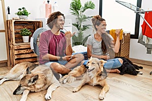 Young hispanic couple doing laundry with dogs smiling with happy face looking and pointing to the side with thumb up