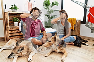 Young hispanic couple doing laundry with dogs smiling and laughing with hand on face covering eyes for surprise