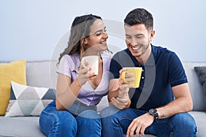 Young hispanic couple drinking coffee sitting on sofa at home