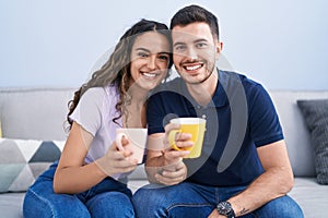 Young hispanic couple drinking coffee sitting on sofa at home