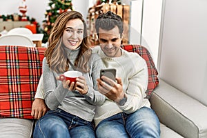 Young hispanic couple drinking coffee using smartphone sitting on the sofa at home