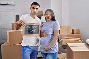 Young hispanic couple expecting a baby moving to a new home puffing cheeks with funny face