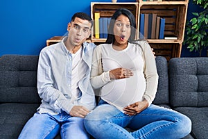 Young hispanic couple expecting a baby sitting on the sofa at home scared and amazed with open mouth for surprise, disbelief face