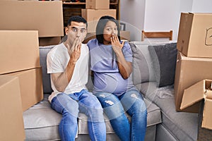 Young hispanic couple expecting a baby sitting on the sofa at new home covering mouth with hand, shocked and afraid for mistake